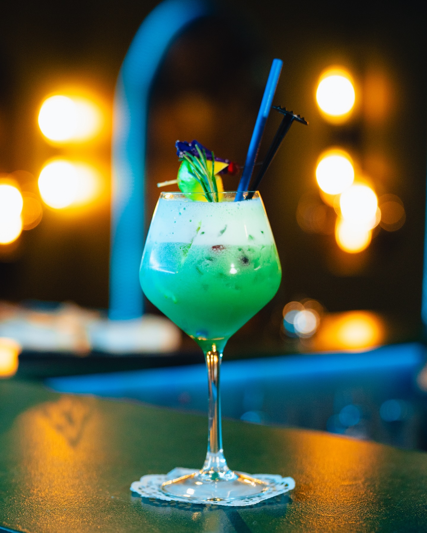 A vibrant green cocktail garnished with mint and edible flowers at a bar.