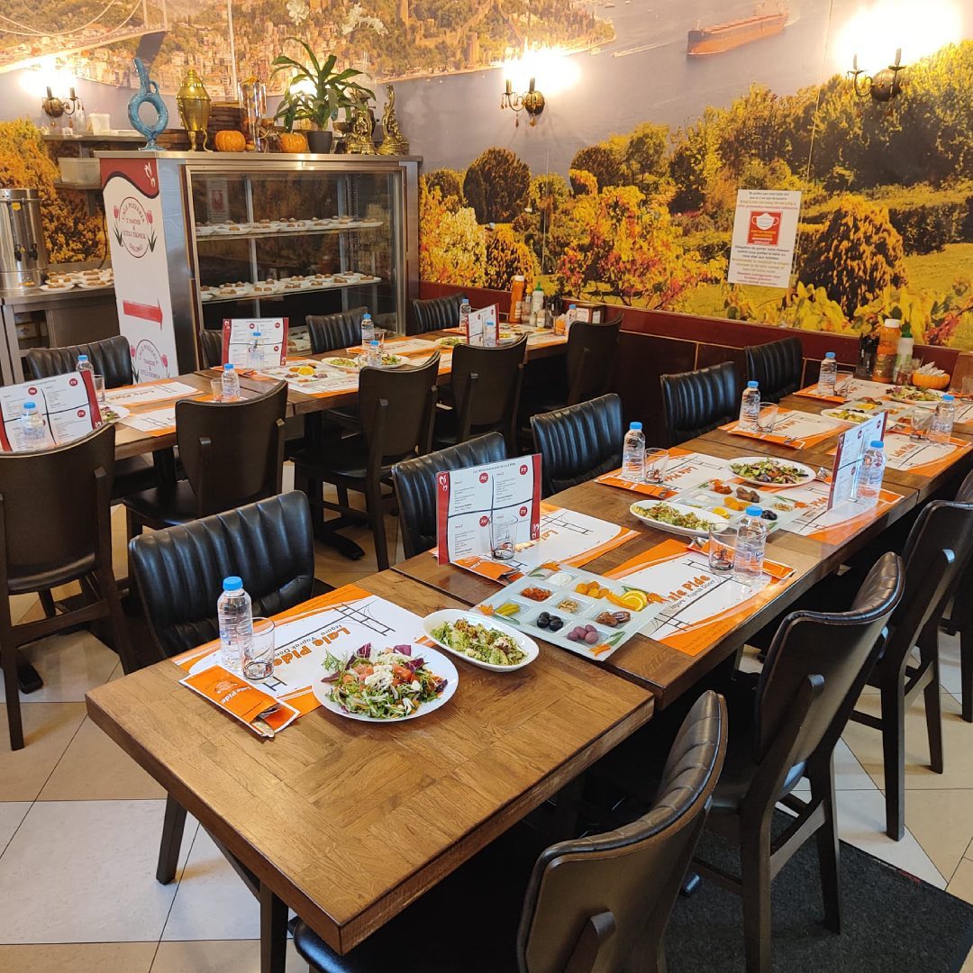 The interior of halal restaurant Lale in Schaerbeek, Belgium, showing wooden tables set with plates of food, menus, and water bottles, with a colorful mural in the background.