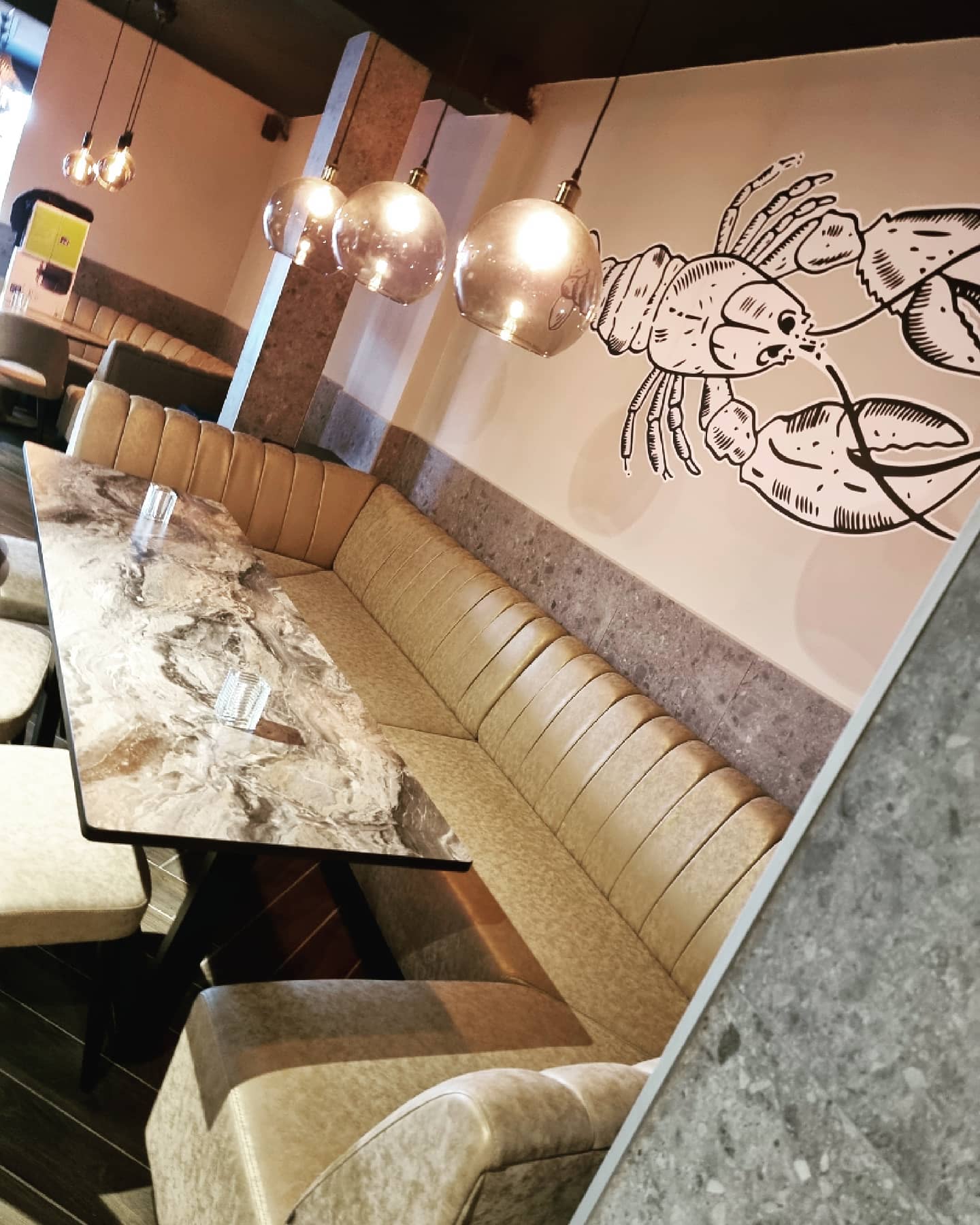 The interior of halal restaurant Zeester Eindhoven in Eindhoven, Netherlands, showing a marble table, beige leather seating, and pendant lighting with a lobster mural on the wall.