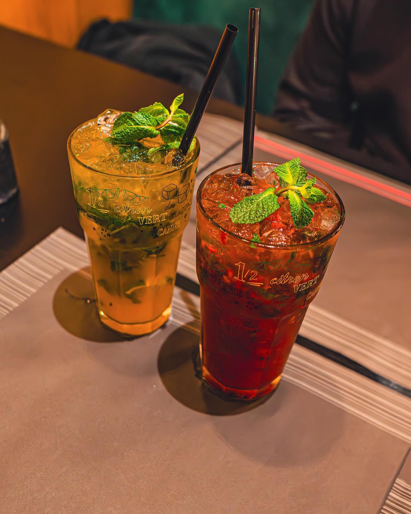Twee drankjes op een tafel in een restaurant, de ene is een groene muntdrank en de andere is een donkerrode drank, beide gegarneerd met muntbladeren.