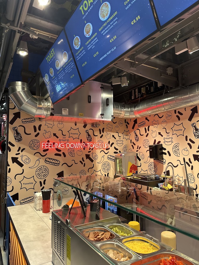 The interior of halal restaurant Toastup in Rotterdam, Netherlands, showing a menu display with various toast options, a food preparation area with ingredients, and a colorful wall design.