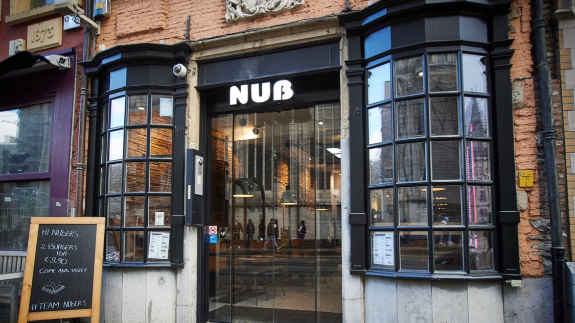 The exterior of halal restaurant Nub Smashburger in Gent, Belgium, showing large glass windows, a black sign with the name 'NUB', and a wooden menu board outside.