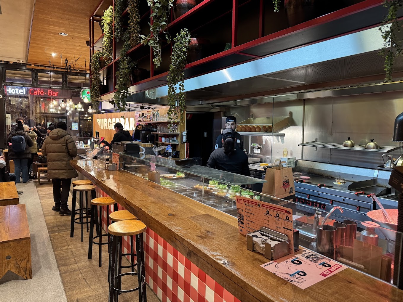 The interior of halal restaurant Burger Bar in Amsterdam, Netherlands, showing a wooden counter, bar stools, a kitchen area with chefs, and a menu on the counter.