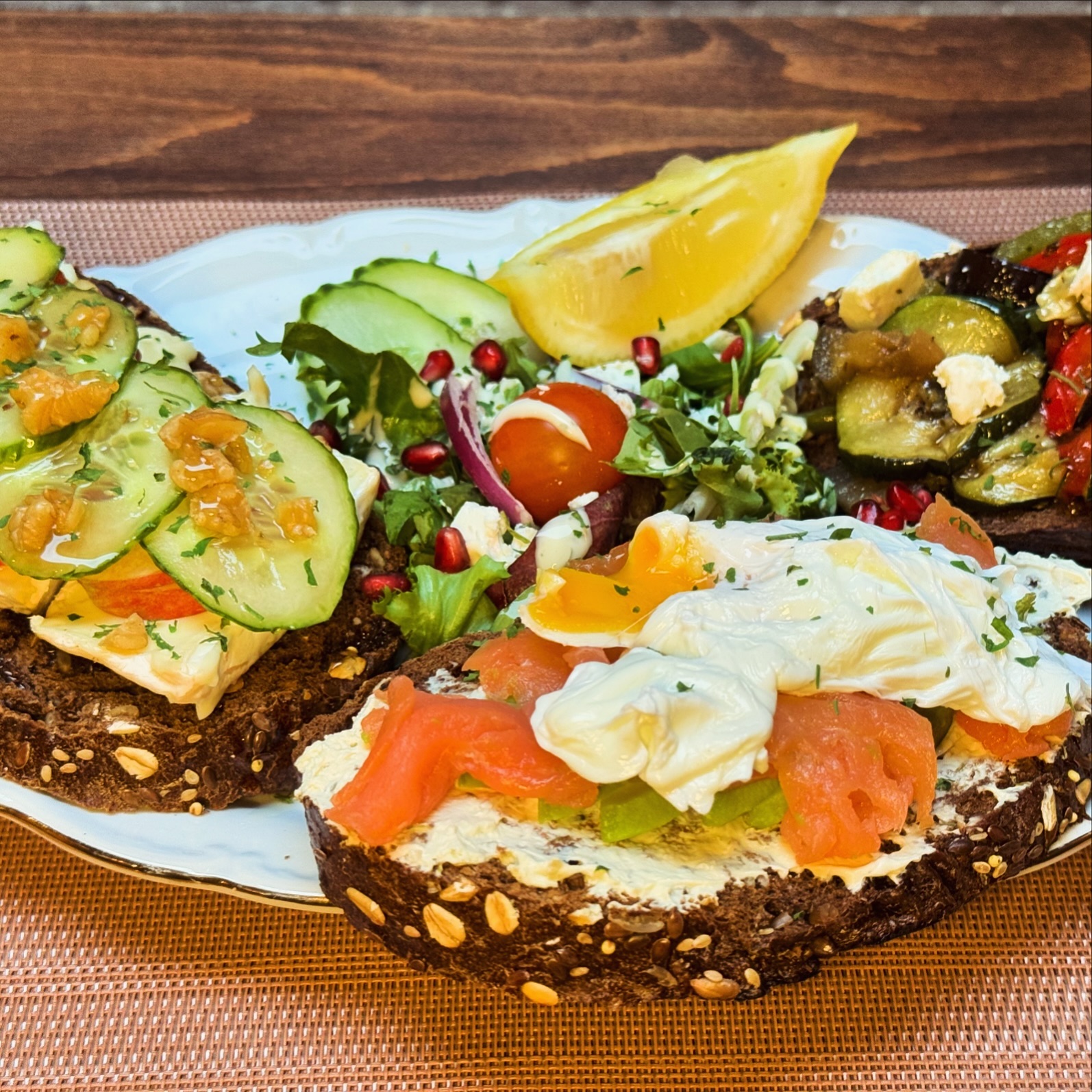 A plate of three open-faced sandwiches featuring various toppings, including cucumbers, tomatoes, and poached eggs, served with a side salad and lemon wedges at O'Tweat, a halal restaurant in Jette, Belgium.