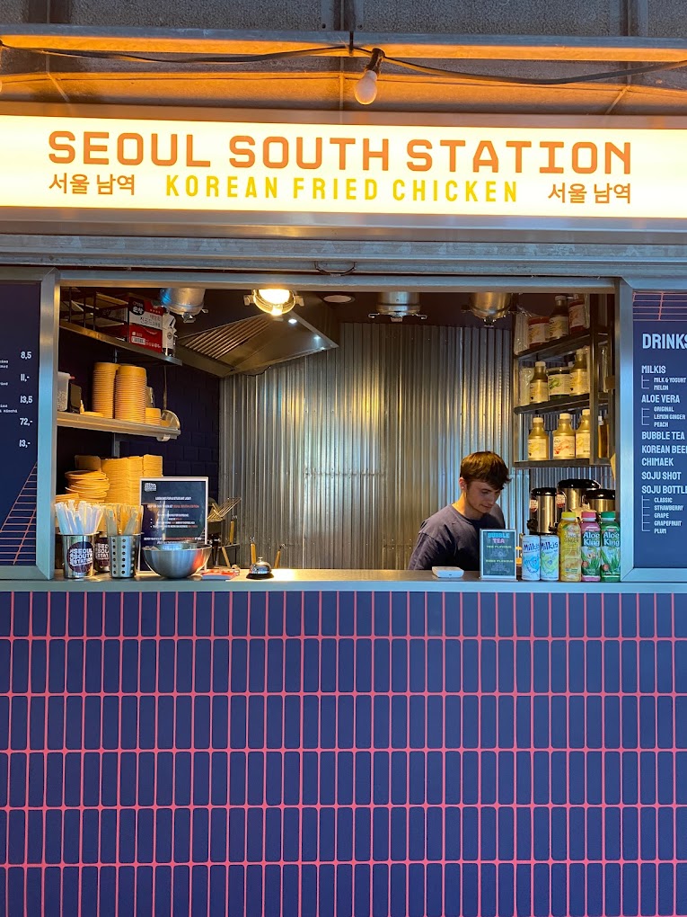 The interior of halal restaurant Seoul South Station in Leuven, Belgium, showing a counter with a staff member, a menu board, and various drinks on display.