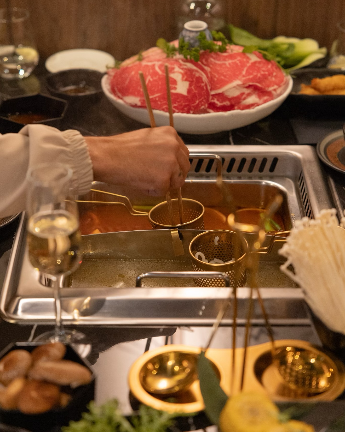 A hot pot dish being prepared at a dining table, featuring a hand using chopsticks to dip ingredients into a pot of broth, with slices of beef and various vegetables in the background at Sorae, a halal restaurant in Eindhoven, Netherlands.