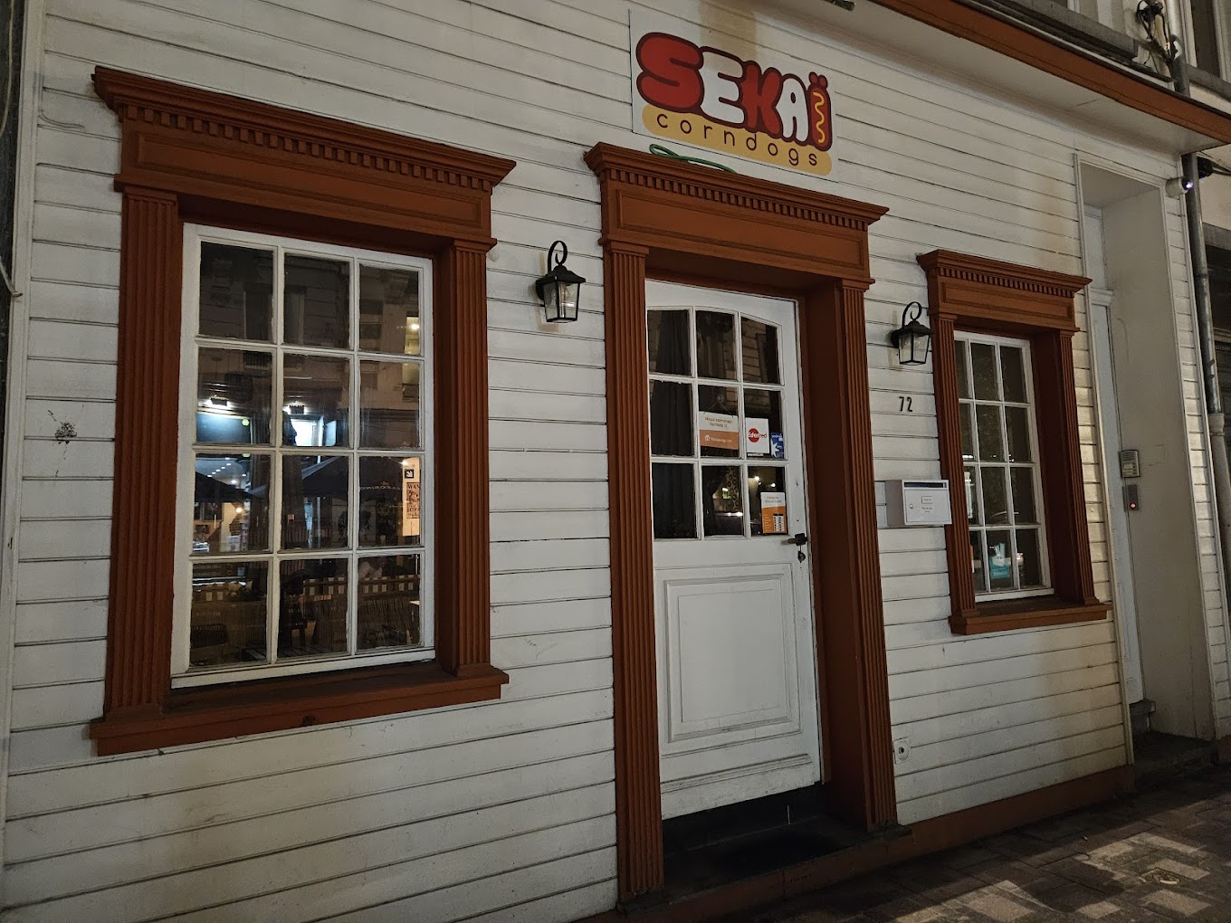 The exterior of halal restaurant Sekaï Corndogs in Bruxelles, Belgium, showing a white wooden facade with orange trim, large windows, and a sign above the door.