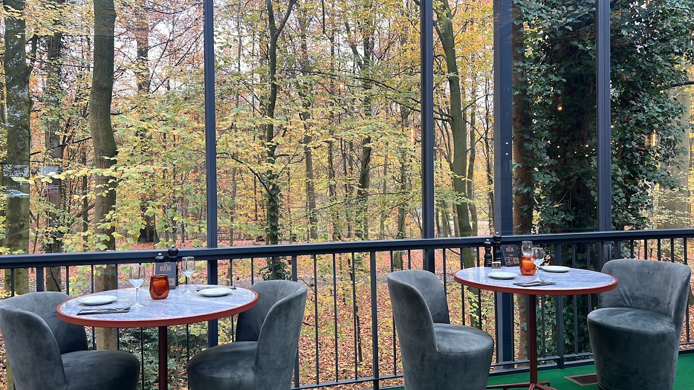 The interior of halal restaurant La Ruche in Bruxelles, Belgium, showing two round tables with marble tops, grey upholstered chairs, and large glass windows overlooking a forest with autumn foliage.