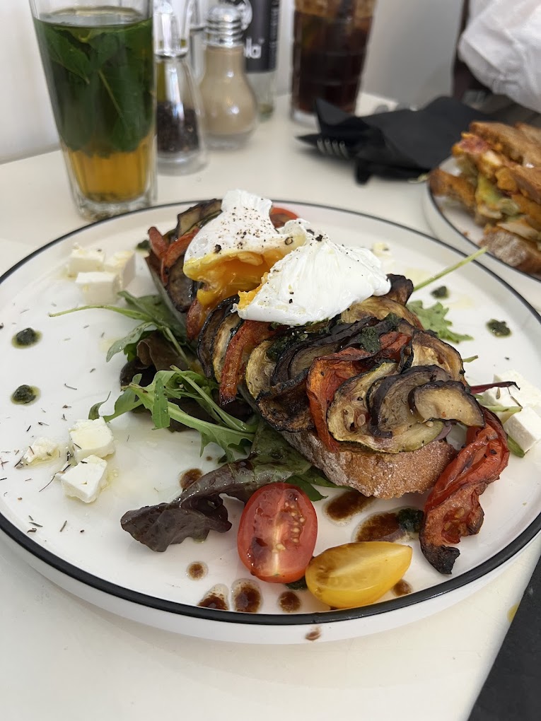 A plate of grilled vegetables on toasted bread topped with poached eggs, served with mixed greens and cherry tomatoes at 2D Café, a halal restaurant in Molenbeek-Saint-Jean, Belgium.