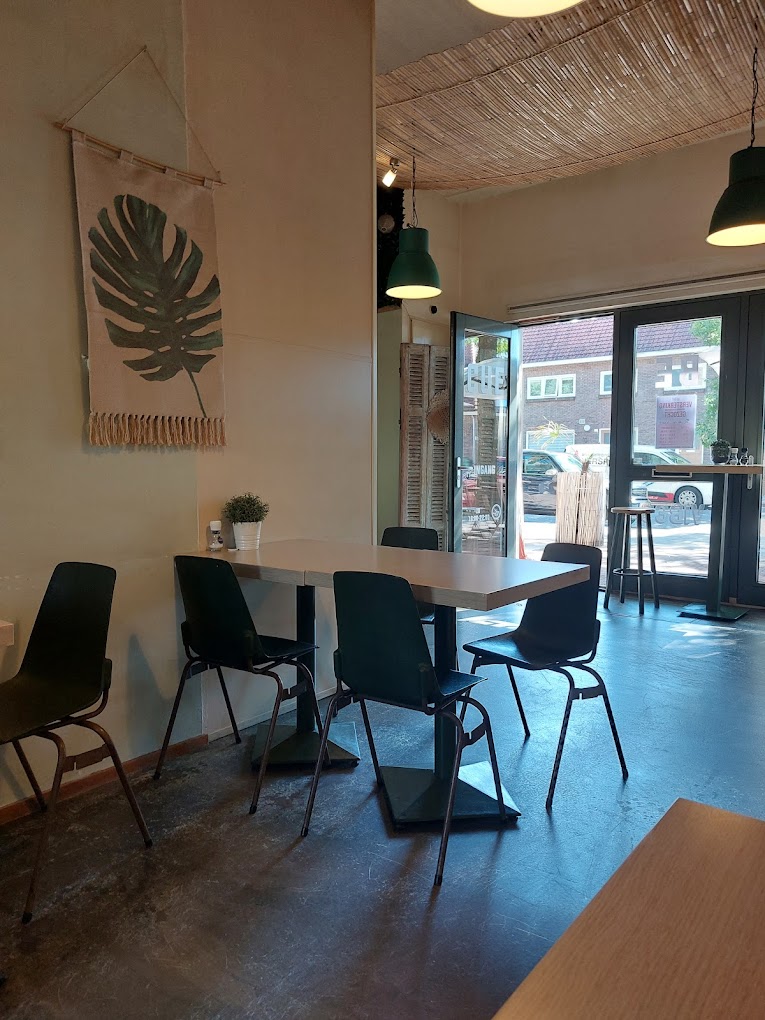 The interior of halal restaurant De Kiet in Eindhoven, Netherlands, showing a minimalist design with a large leaf wall hanging, green pendant lights, and empty black chairs around a white table.