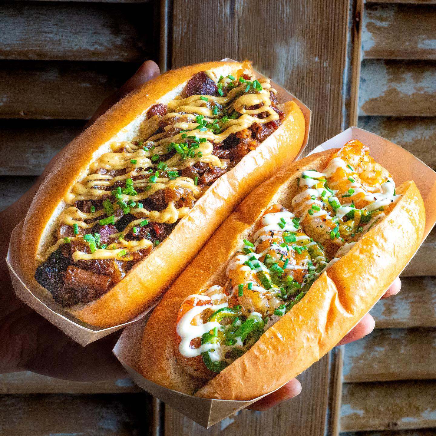 A close-up of two gourmet hot dogs at a restaurant, one topped with grilled meat and drizzled with sauce, and the other filled with shrimp and garnished with mayonnaise and herbs.
