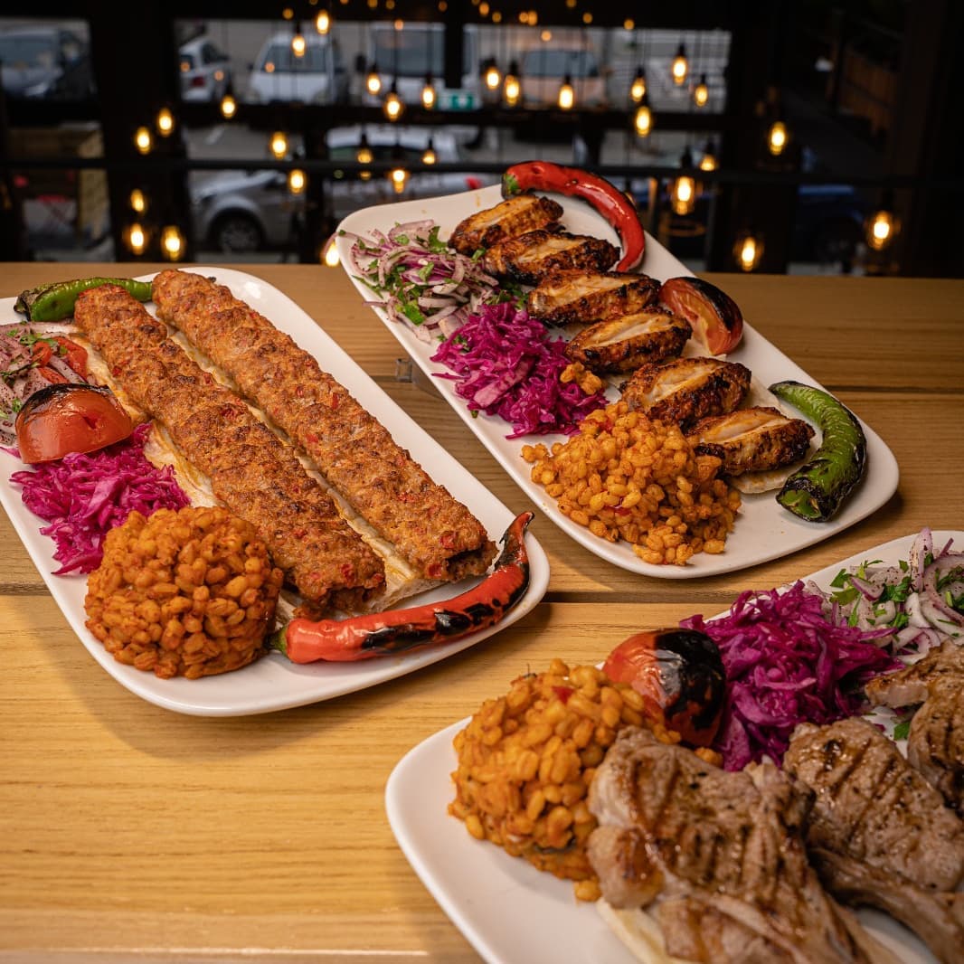 A close-up view of three plates of halal food at Ganii Kebap in Rotterdam, Netherlands. The plates feature various grilled meats, including kebabs, chicken, and lamb chops, served with bulgur wheat and red cabbage salad.