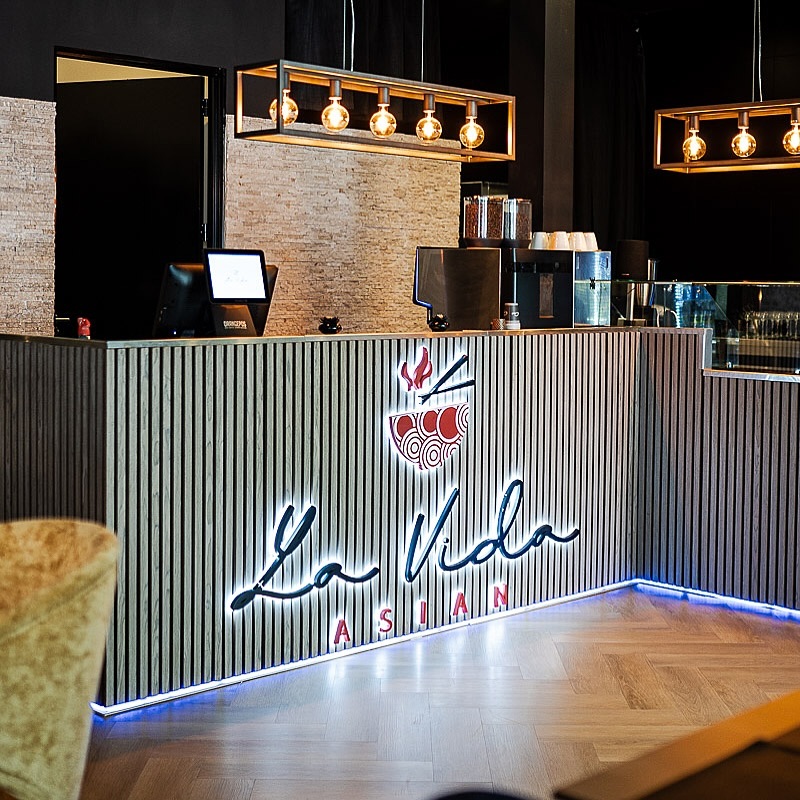 The interior of halal restaurant La Vida in Utrecht, Netherlands, showing a modern reception area with wooden paneling, a lit sign, and stylish pendant lights.
