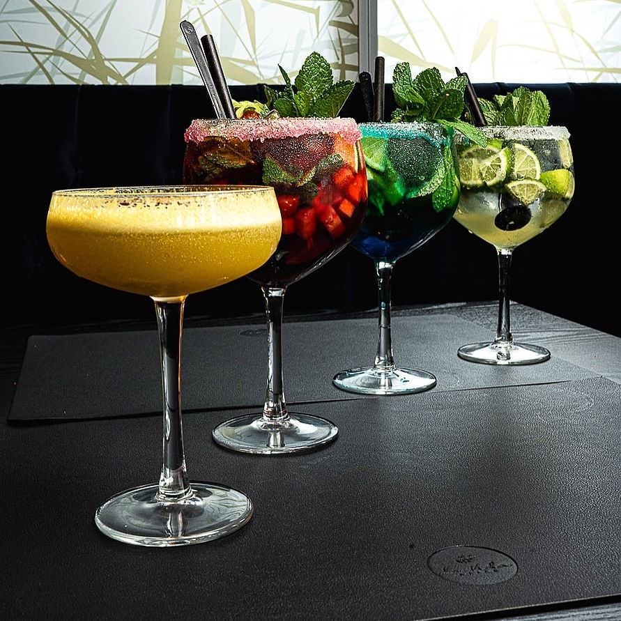 A selection of colorful cocktails at a bar, featuring a yellow drink, a fruit-filled drink with mint, and two other vibrant drinks with garnishes.