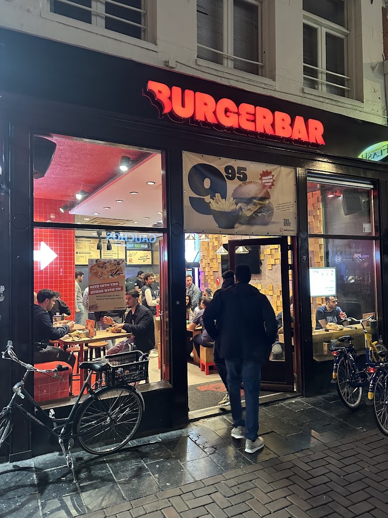 The exterior of halal restaurant Burger Bar in Amsterdam, Netherlands, showing a brightly lit sign and people dining inside.