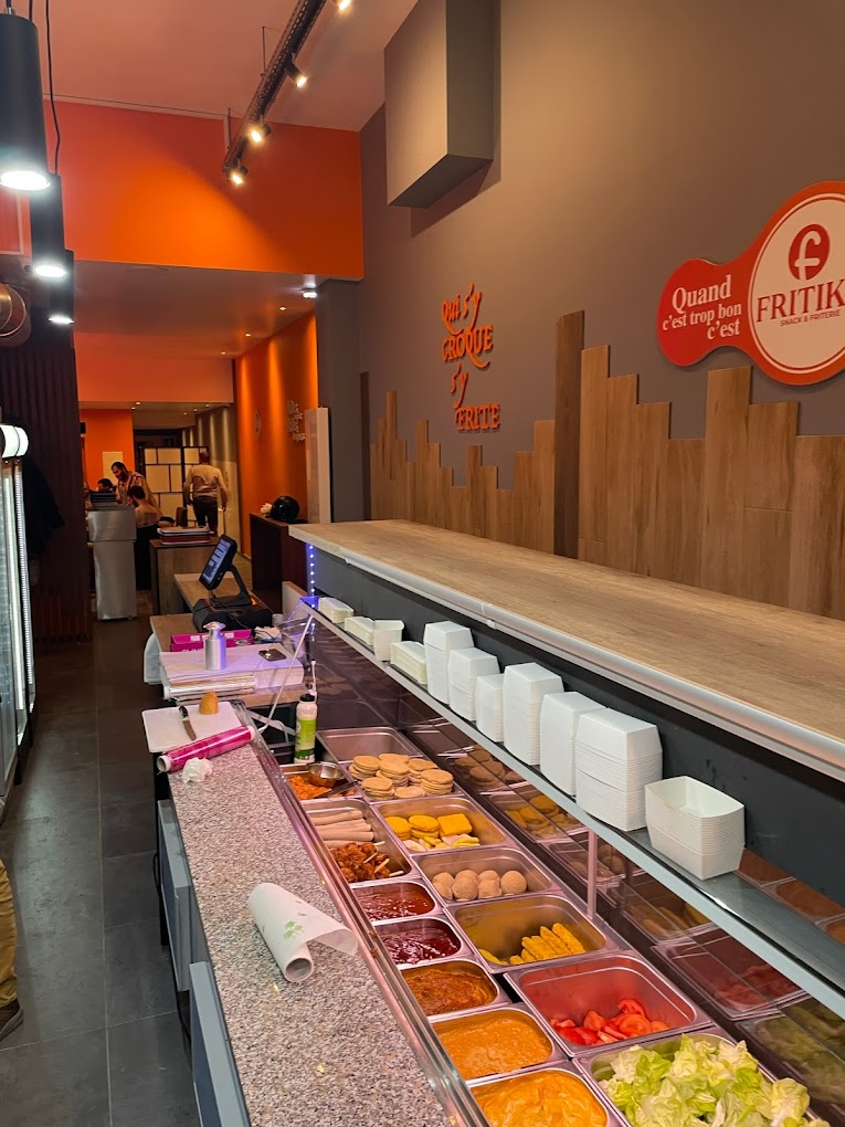 L'intérieur du restaurant halal Fritiko à Anderlecht, en Belgique, montrant un comptoir avec divers aliments, y compris des sauces, des légumes et des collations, ainsi qu'un design moderne avec des accents orange.