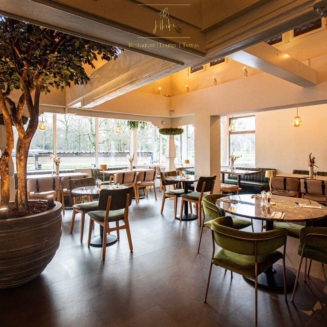 The interior of Hkiki in Rotterdam, Netherlands, showing modern furniture, large windows, and indoor trees.