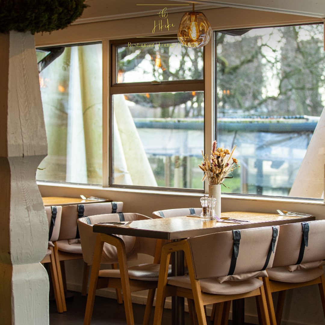 The interior of Hkiki in Rotterdam, Netherlands, showing a cozy dining area with wooden tables, beige upholstered chairs, and large windows allowing natural light.