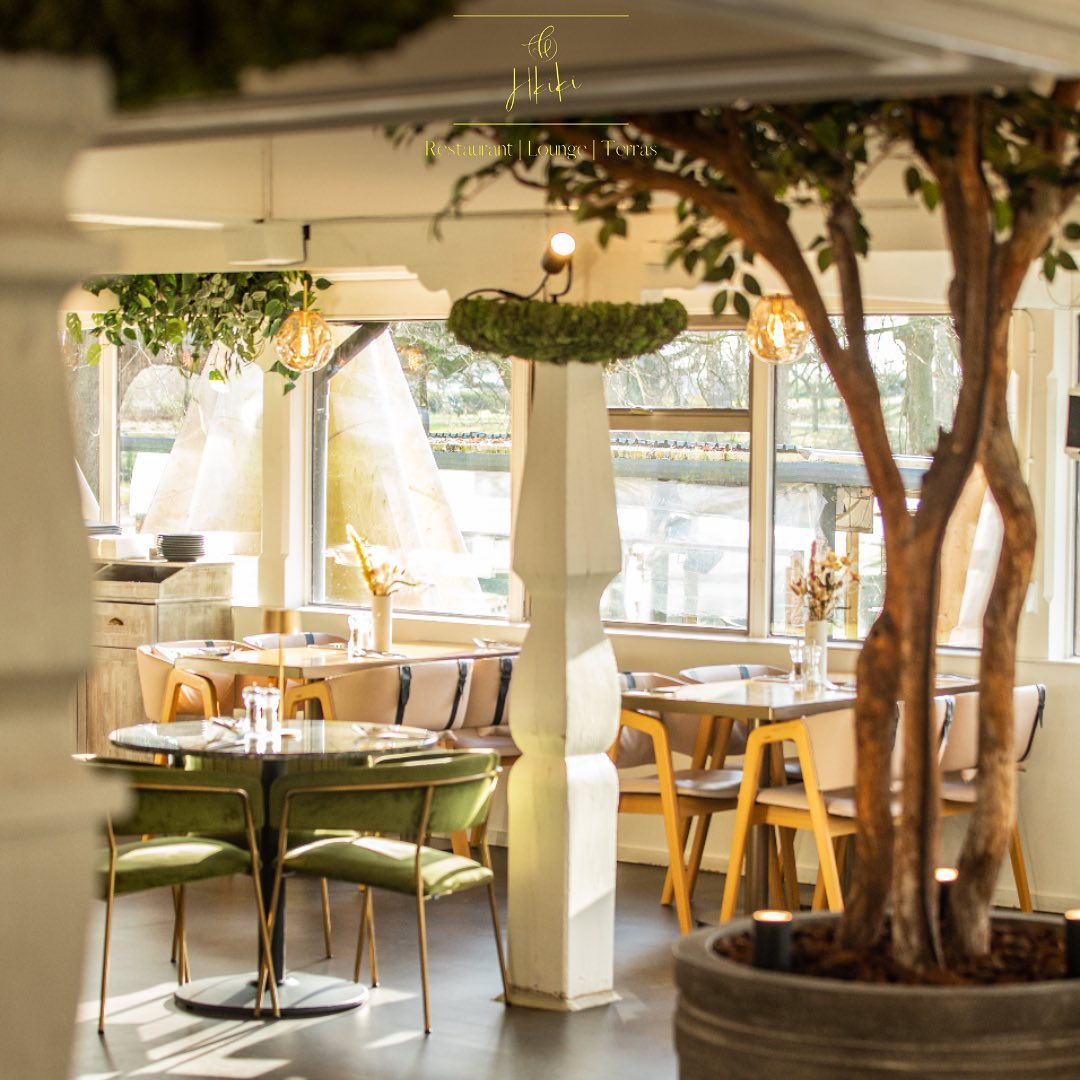 The interior of Hkiki in Rotterdam, Netherlands, showing a modern dining area with green and wooden furniture, large windows, and decorative plants.