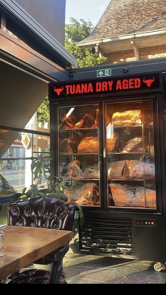 The exterior of halal restaurant Tuana in Sint-Niklaas, Belgium, showing a dry aging meat display with various cuts of meat visible inside a glass refrigerator.