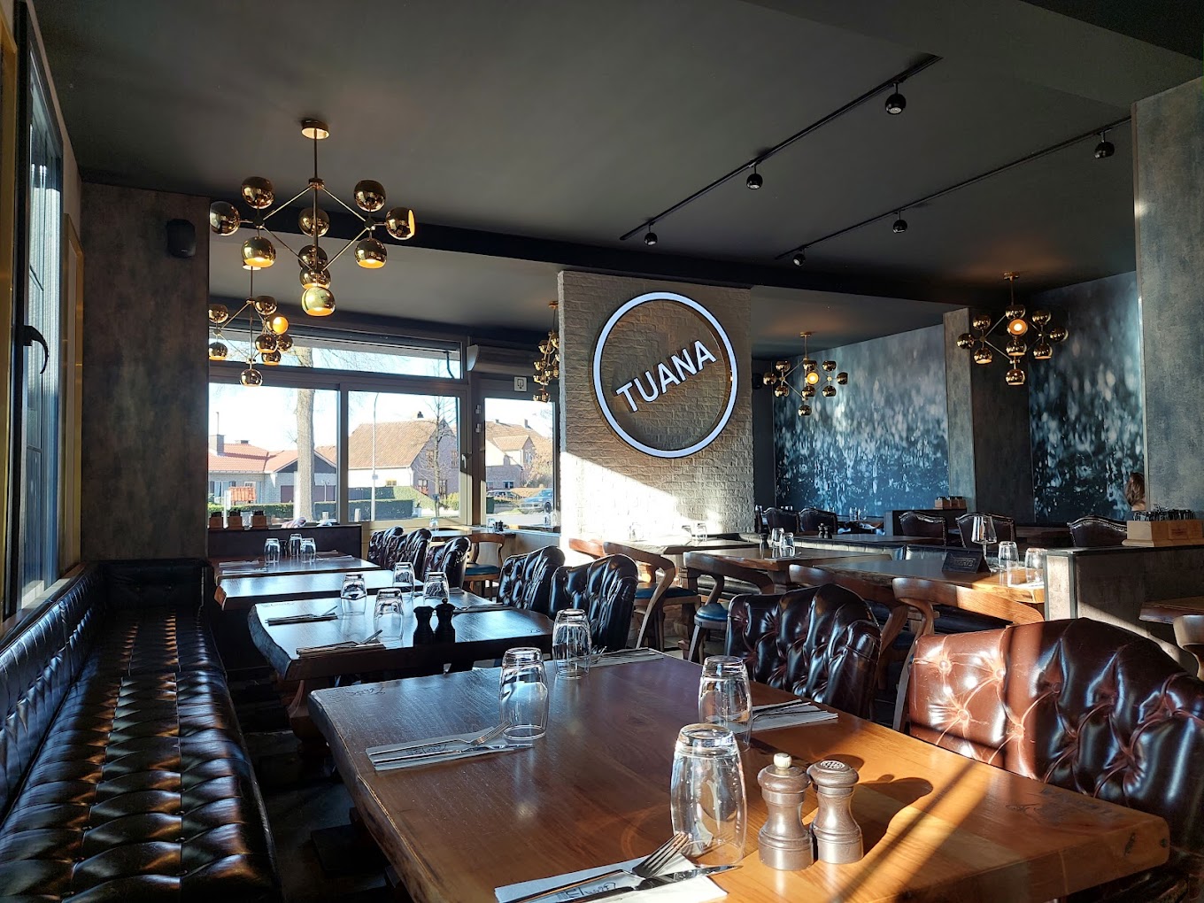 The interior of halal restaurant Tuana in Sint-Niklaas, Belgium, showing modern decor, wooden tables, leather seating, and large windows with natural light.