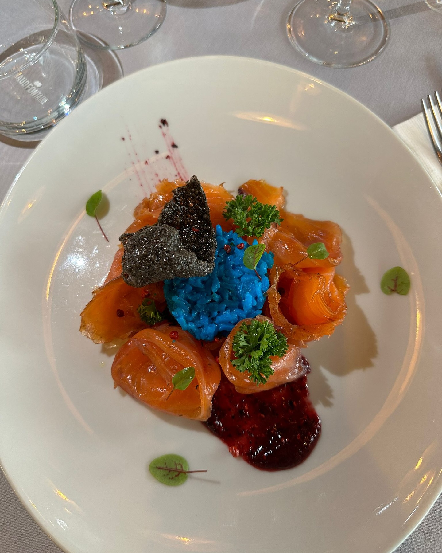Een bord gerookte zalm met blauwe risotto, gegarneerd met kruiden en een donker krokant element in halal restaurant The Manzoni in Brussel, België.