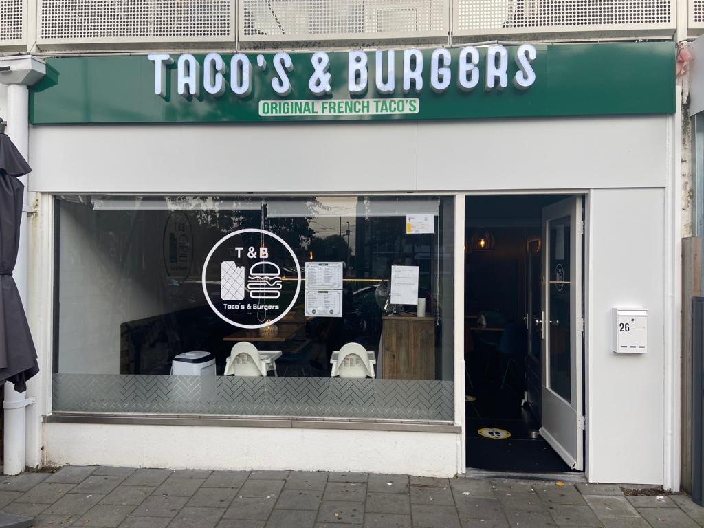 The exterior of halal restaurant Taco's and Burgers in Amsterdam, Netherlands, showing the restaurant's sign and large windows with a view of the interior seating area.