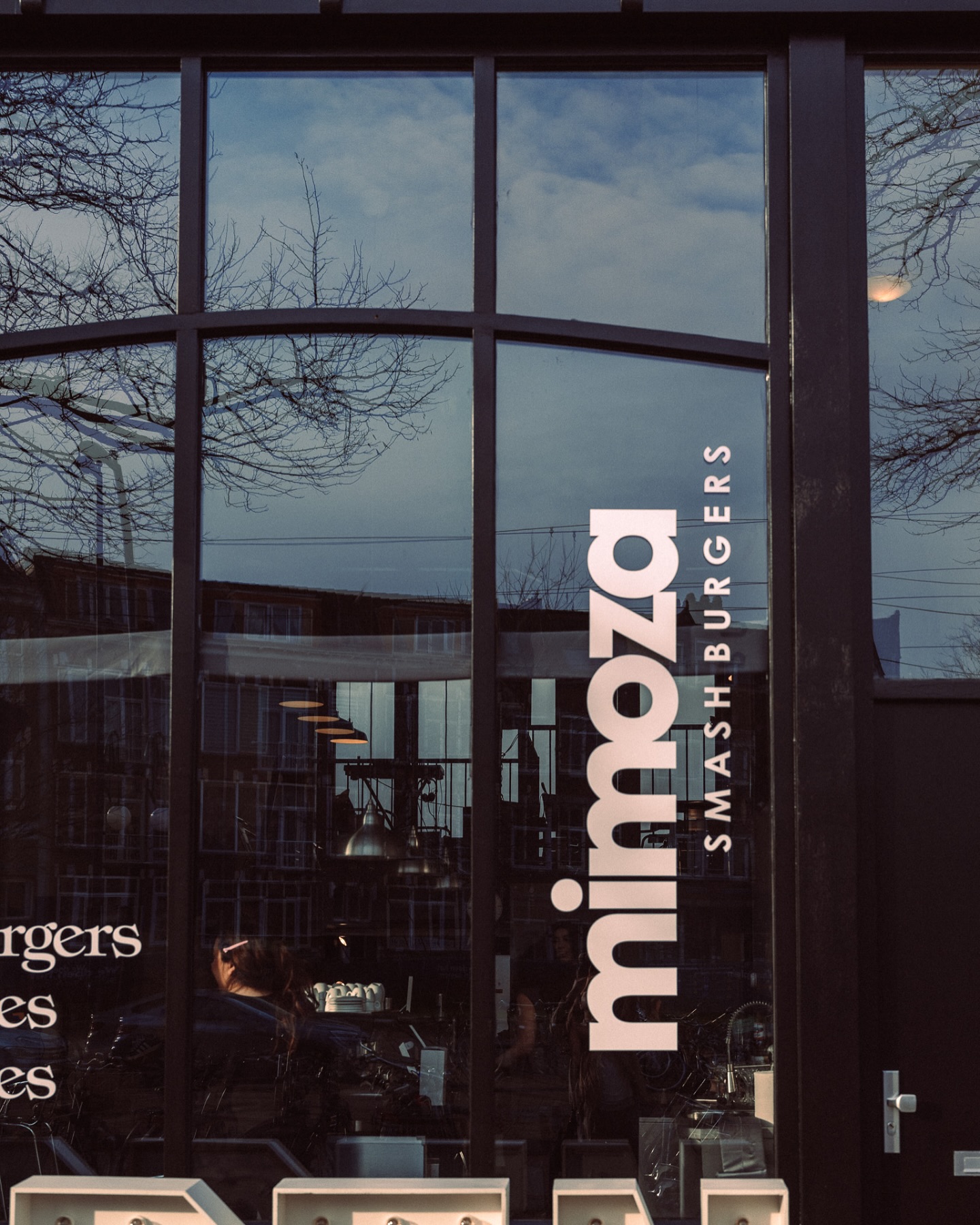 The exterior of halal restaurant Mimoza Smashburgers in Rotterdam, Netherlands, showing large glass windows with reflections and a prominent sign displaying the restaurant's name.