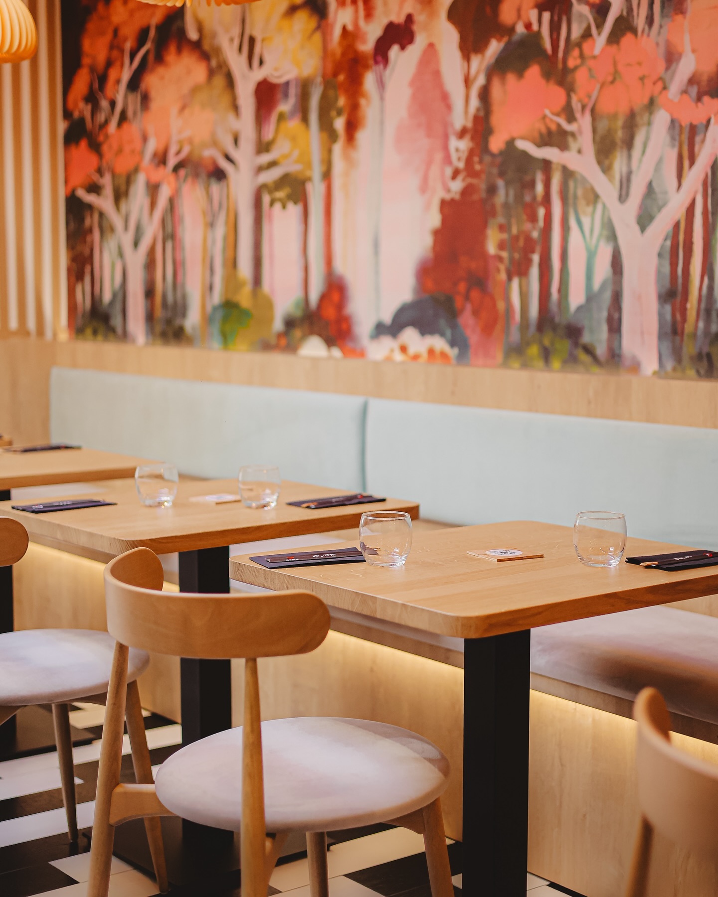 Het interieur van halal restaurant Tsuki in Anderlecht, België, met houten tafels, lichte stoelen en een kleurrijke muurschildering van bomen.