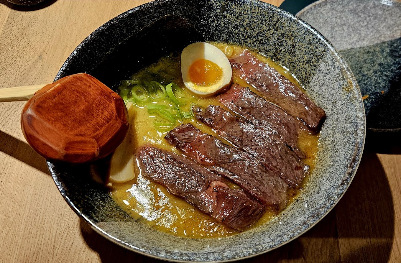 A bowl of ramen with slices of beef, a soft-boiled egg, green onions, and a wooden ladle at Maruki, a halal restaurant in Rotterdam, Netherlands.