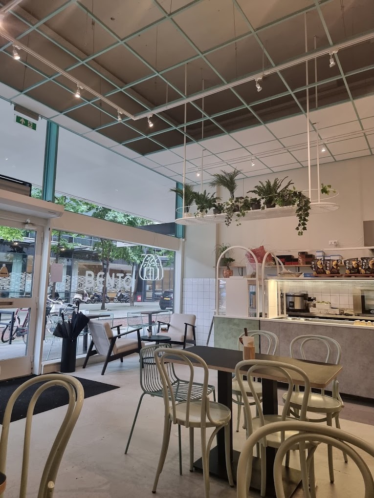 The interior of halal restaurant Bento Café in Rotterdam, Netherlands, showing a modern design with hanging plants, a light color scheme, and seating arrangements including chairs and tables.