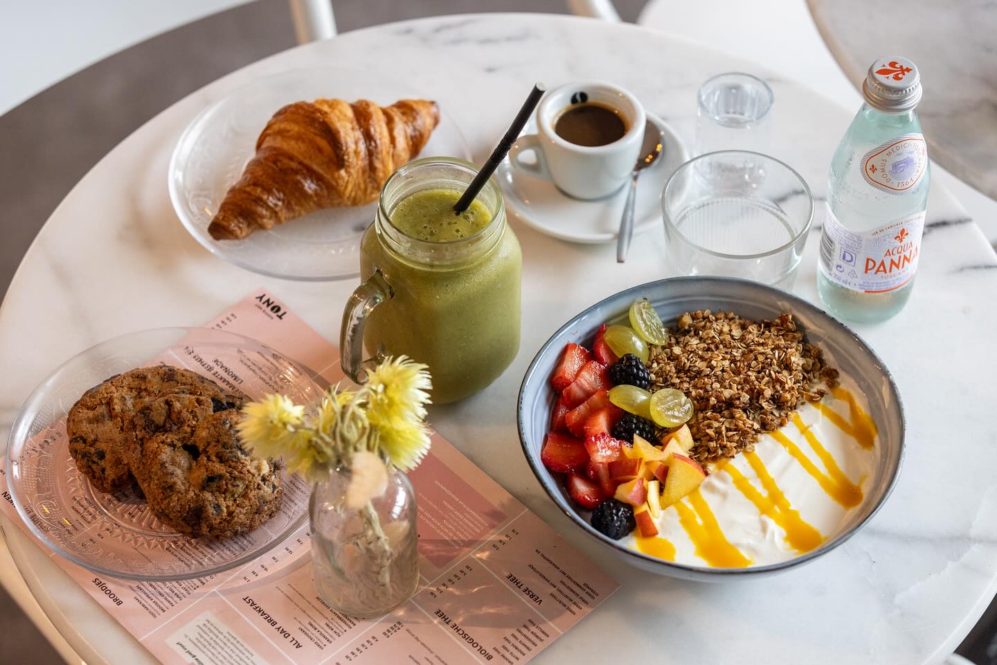 A breakfast spread featuring a croissant, a green smoothie, a bowl of yogurt topped with granola and fresh fruits, two cookies, and a cup of espresso at Koffie Met Tony, a halal restaurant in Ede, Netherlands.