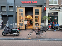 The exterior of halal restaurant Burger Bar in Amsterdam, Netherlands, showing large wooden doors and outdoor seating with patrons.