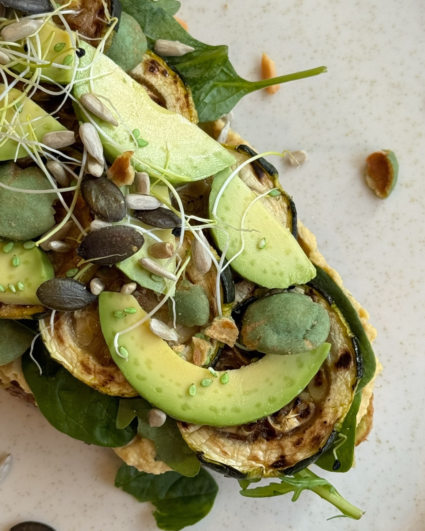 A colorful open sandwich topped with grilled zucchini, avocado slices, spinach, and various seeds at The Bread Bar, a halal restaurant in Gent, Belgium.
