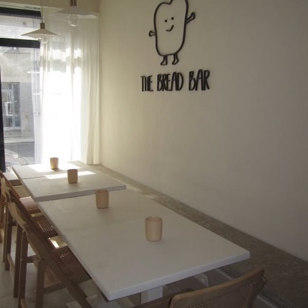 The interior of halal restaurant The Bread Bar in Gent, Belgium, showing white tables, wooden chairs, and a wall with a cartoon bread character and the restaurant's name.