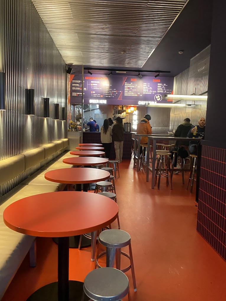 The interior of halal restaurant Seoul South Station in Gent, Belgium, showing red tables, metal stools, and a menu board on the wall.