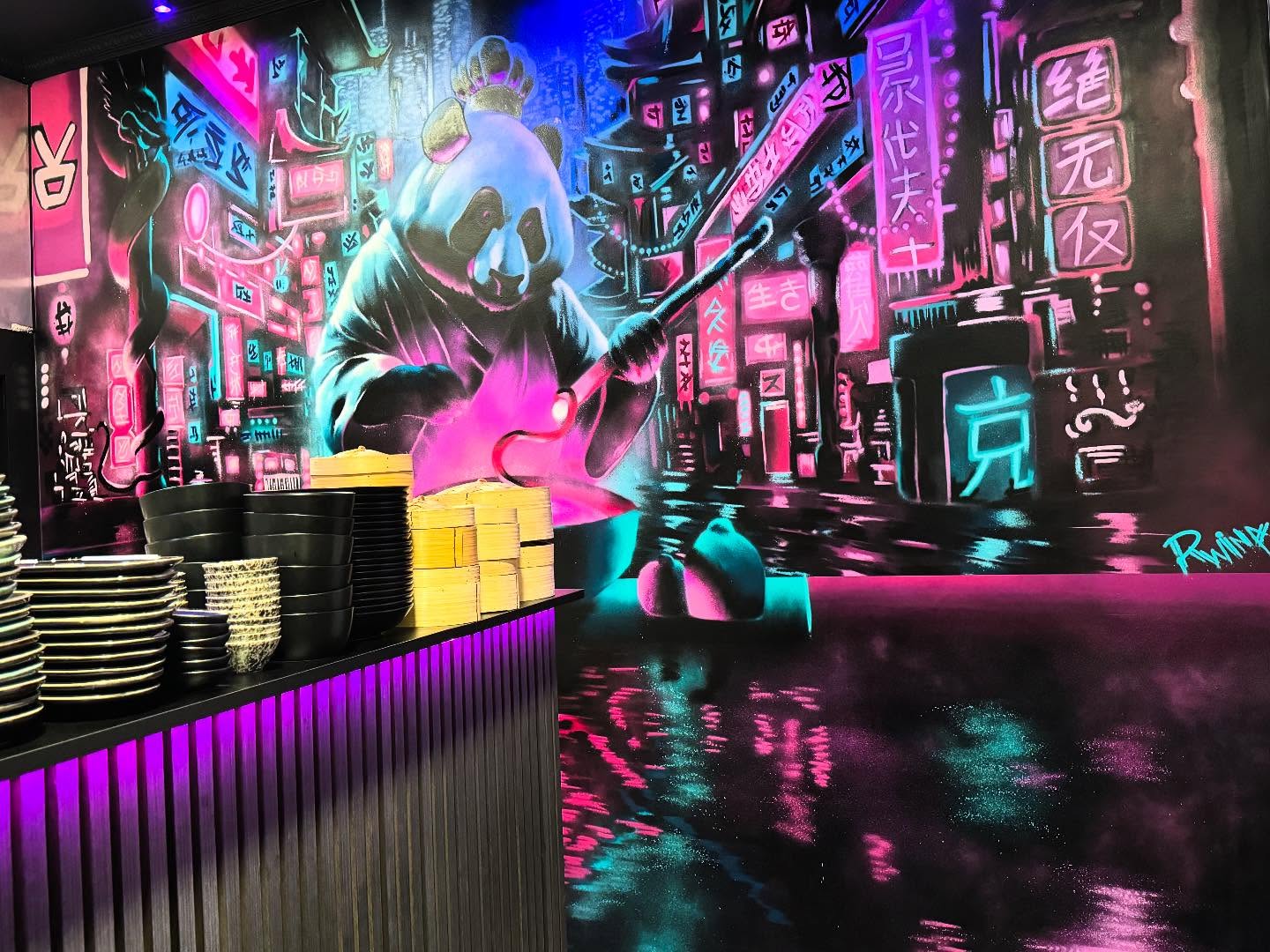 The interior of halal restaurant Wokcity 'N Roll in Mortsel, Belgium, showing a vibrant mural of a panda in a neon-lit cityscape, with black plates stacked on a counter and a purple-lit bar.