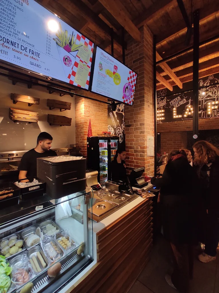 The interior of halal restaurant Joe La Frite in Bruxelles, Belgium, showing a counter with a menu display, a staff member taking orders, and customers waiting in line.