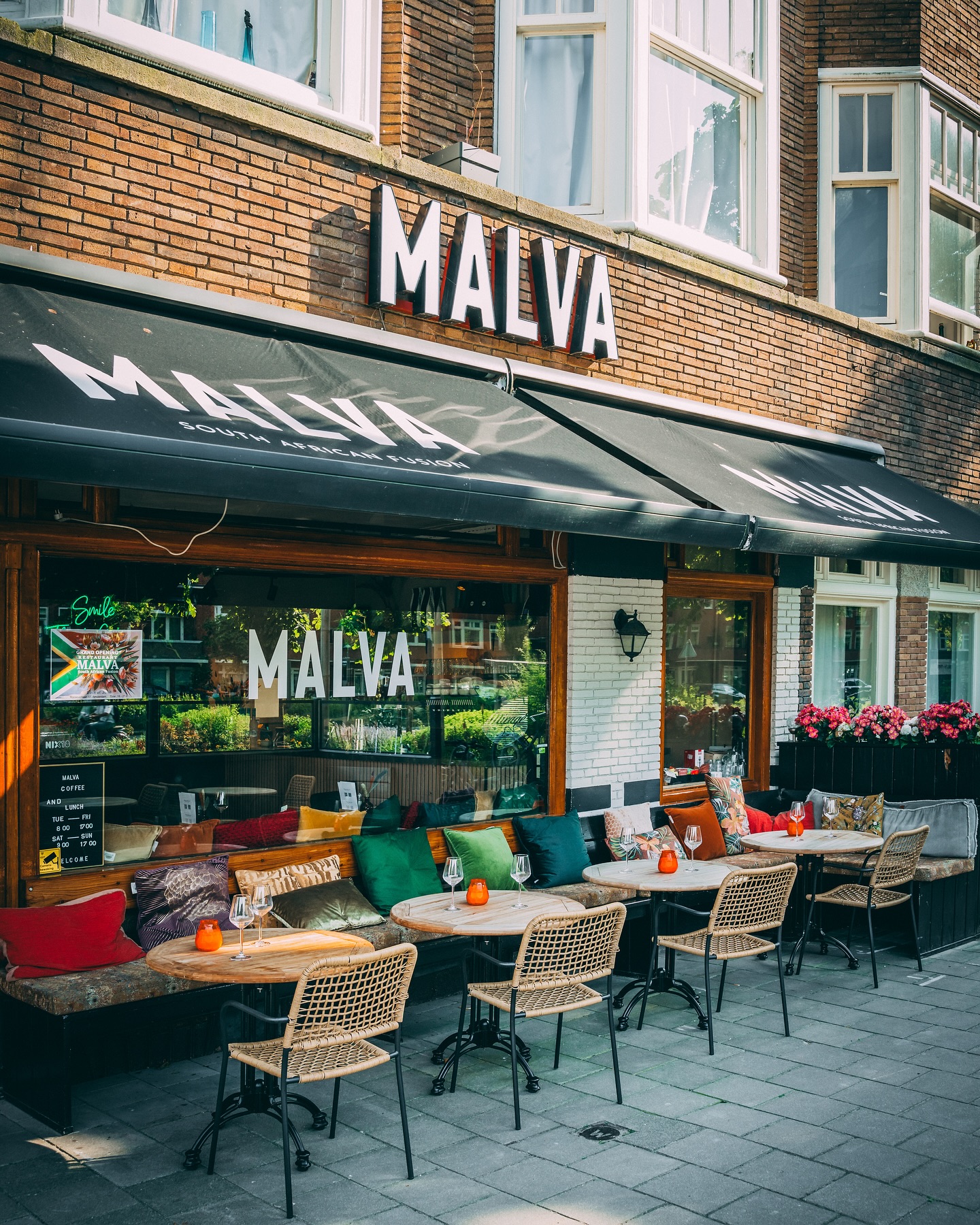The exterior of halal restaurant Malva in Amsterdam, Netherlands, showing a brick facade, large windows, outdoor seating with colorful cushions, and a sign with the restaurant's name.