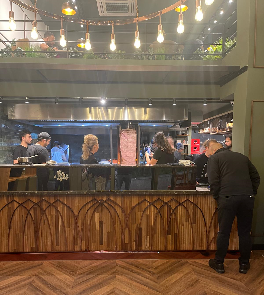The interior of halal restaurant Harbii in Rotterdam, Netherlands, showing a modern design with a wooden bar, hanging lights, and staff preparing food behind the counter.