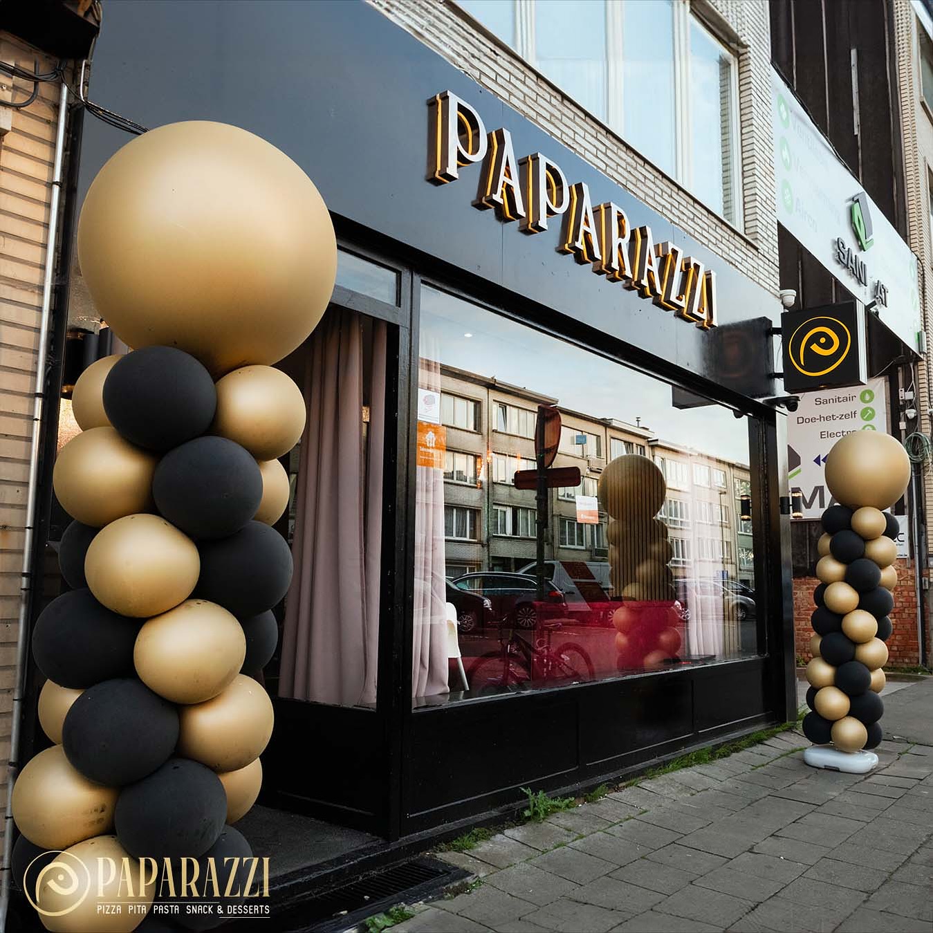 The exterior of halal restaurant Paparazzi Bistro in Antwerpen, Belgium, showing a black and gold color scheme with balloon decorations and large windows.