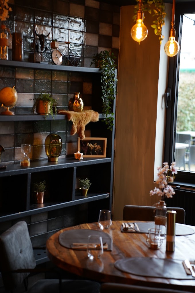 The interior of halal restaurant Buffalo in Beringen, Belgium, showing a wooden table set with glassware and cutlery, decorative shelves with plants and various ornaments, and warm lighting from hanging bulbs.