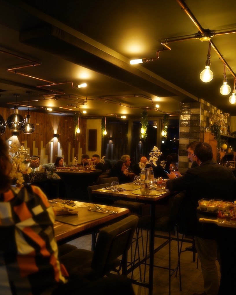 The interior of halal restaurant Buffalo in Beringen, Belgium, showing a modern design with wooden accents, copper piping, and dim lighting, with patrons seated at tables and a bar area.