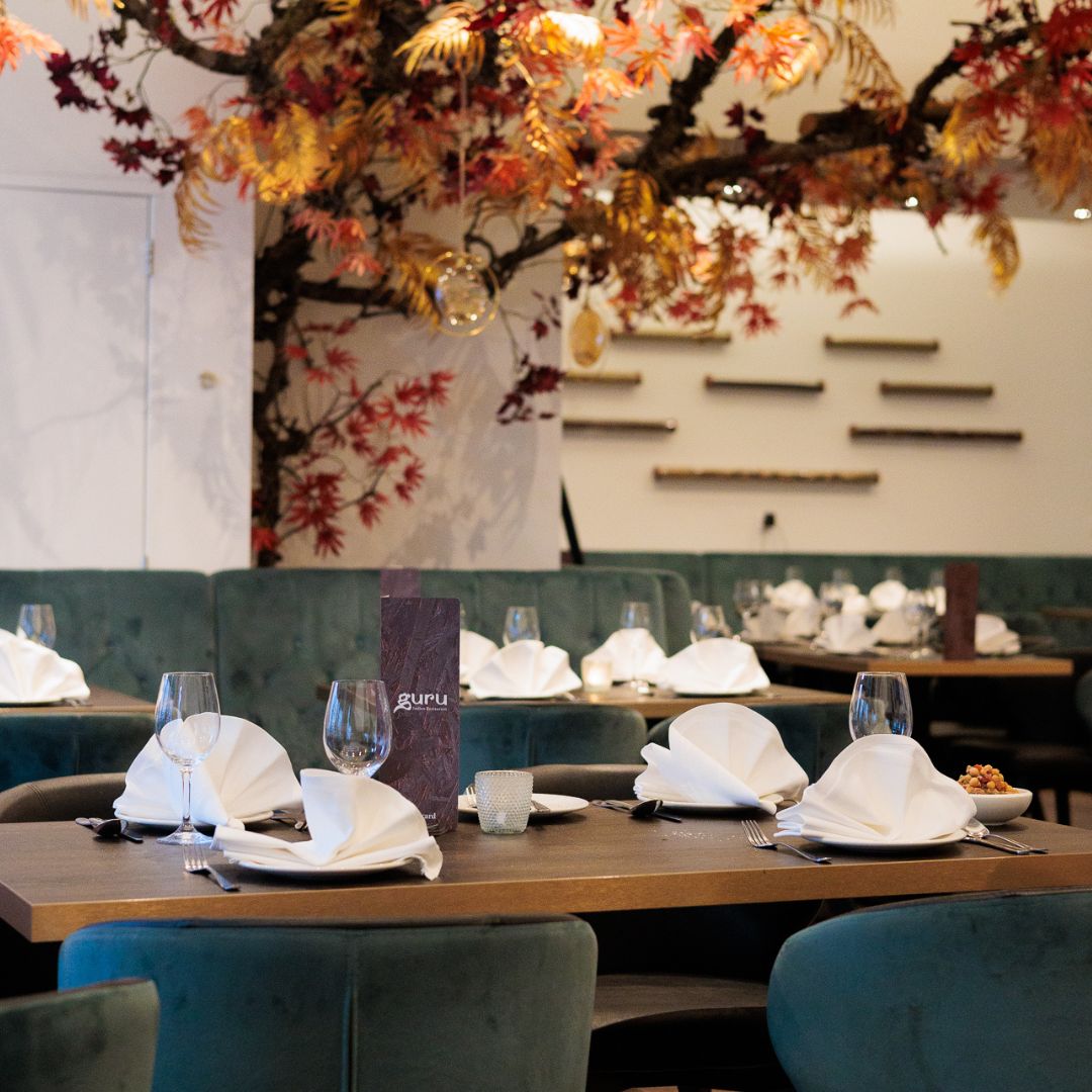 The interior of halal restaurant Guru in Amsterdam, Netherlands, showing elegant green seating, neatly arranged tables with white napkins, and decorative autumn leaves overhead.
