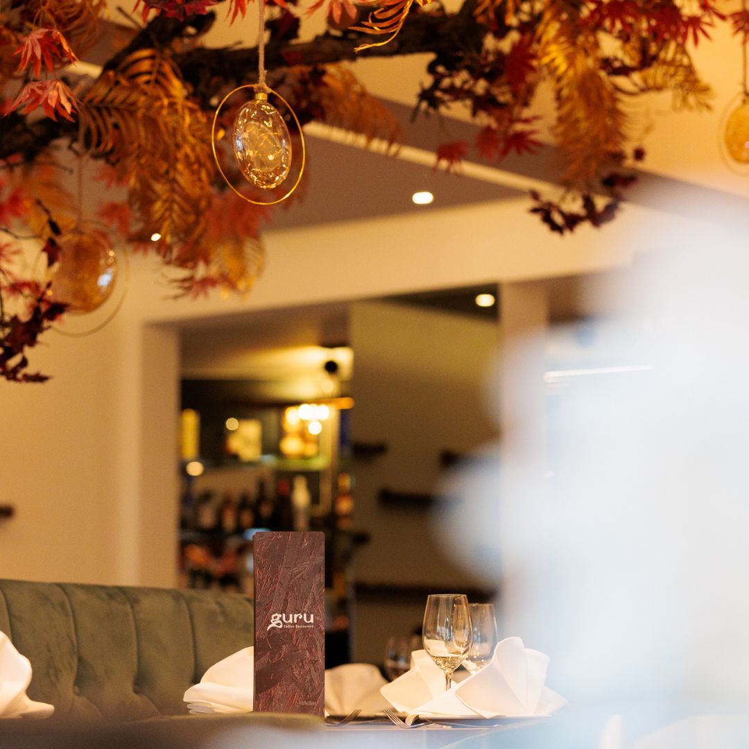 The interior of halal restaurant Guru in Amsterdam, Netherlands, showing a decorated ceiling with hanging glass ornaments and a wooden menu stand on a table with folded napkins and wine glasses.