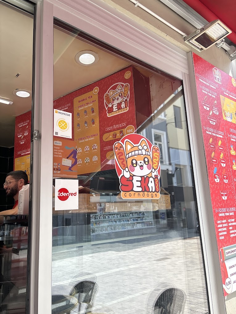 The exterior of halal restaurant Sekaï Corndogs in Bruxelles, Belgium, showing a large logo on the window and colorful menu posters inside.