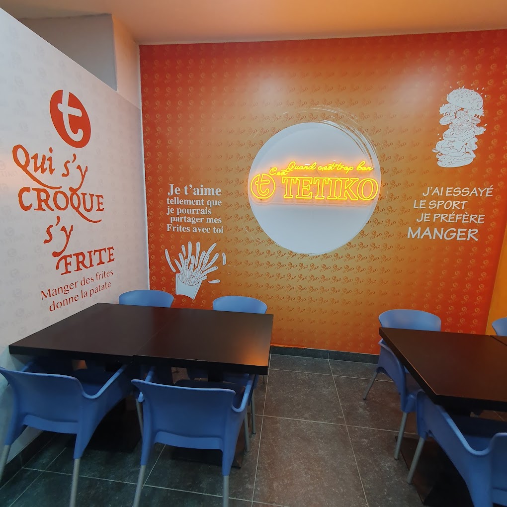 L'intérieur du restaurant halal Fritiko à Ganshoren, en Belgique, montrant des murs orange avec des textes ludiques, une enseigne au néon et des chaises bleues autour de tables noires.