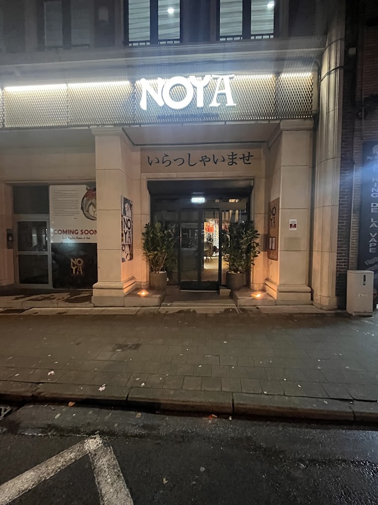 De buitenkant van halal restaurant NOYA in Schaerbeek, België, met de ingang waarop de naam 'NOYA' verlicht is en Japanse tekst boven de deur.
