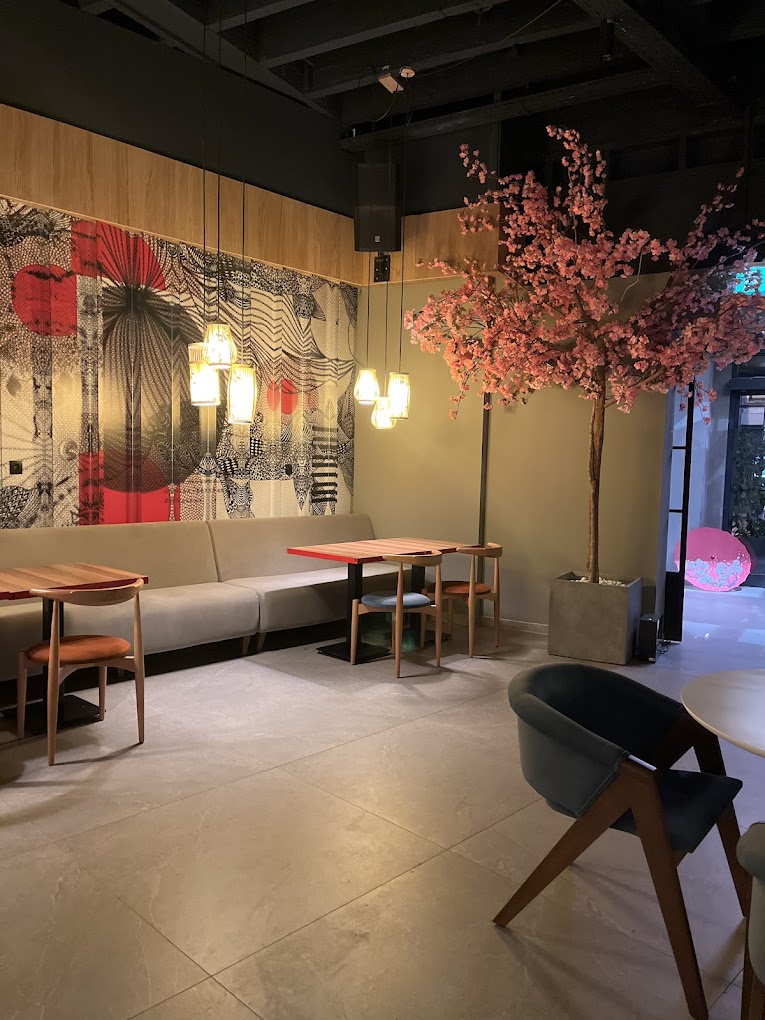 Het interieur van halal restaurant NOYA in Schaerbeek, België, met een modern ontwerp met een grote muurschildering, een roze kersenbloesemboom en hanglampen boven houten tafels.