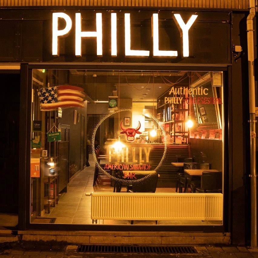 The exterior of halal restaurant Philly Sandwich Bar in Antwerpen, Belgium, showing illuminated signage and a large window displaying the interior.